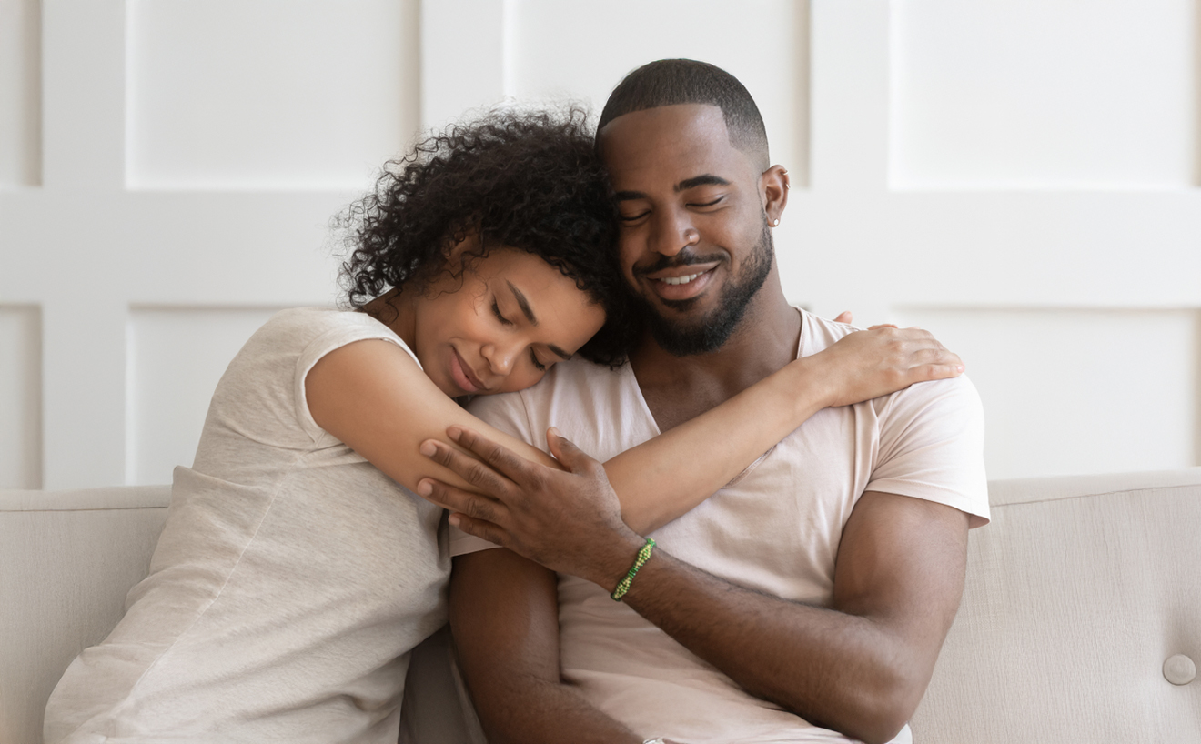 A black couple with their eyes closed and smiling sweetly embracing each other on a couch.